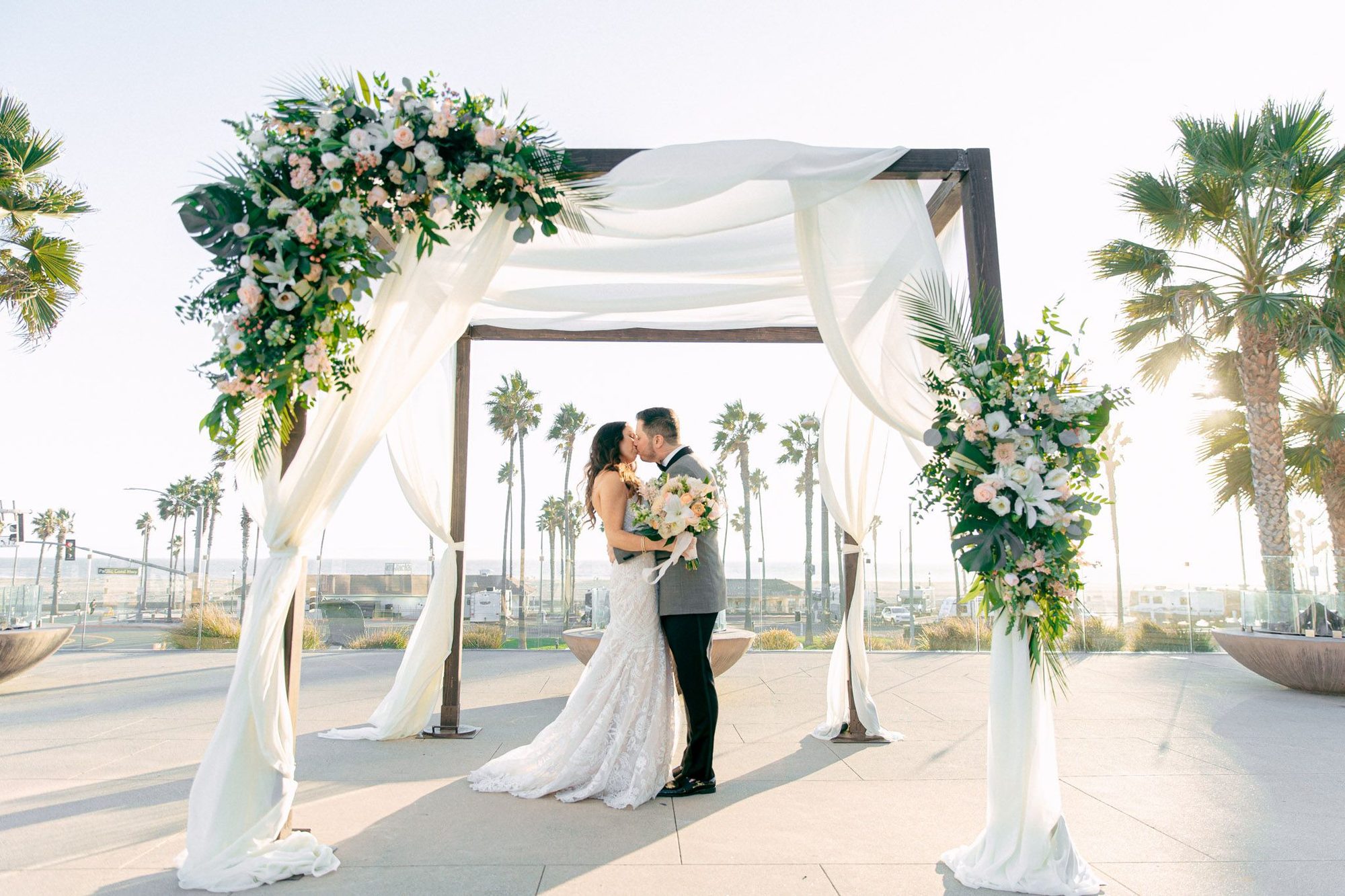 Ceremony arch with white drapes and palms, the couple kissing