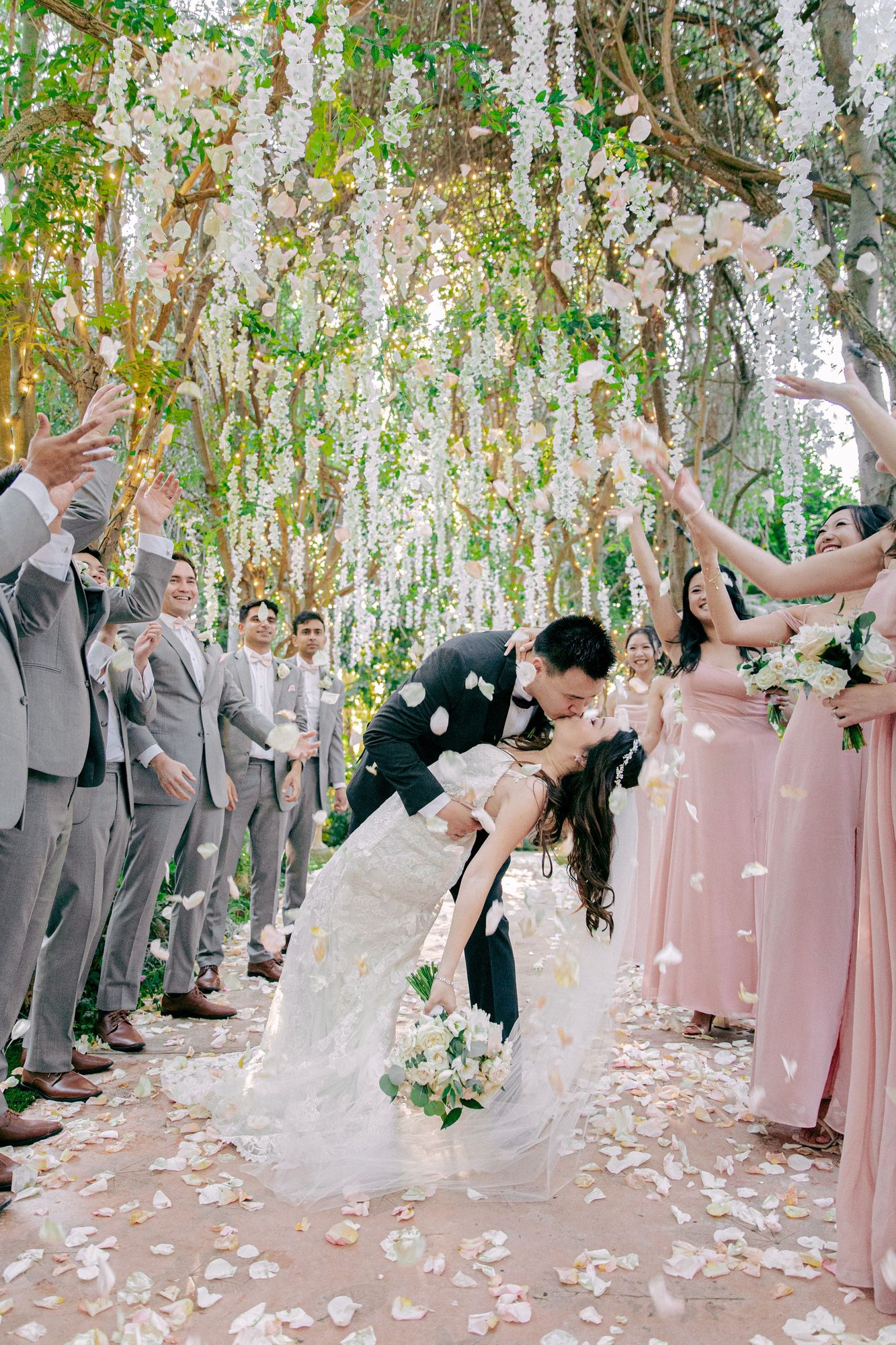Groom dips the bride under hanging flowers as the bridal party cheers